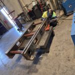 Worker and fabricated steel members on a welding fixture or table. View 1 of 2