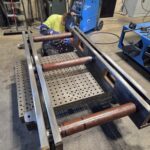 Worker and fabricated steel members on a welding fixture or table. View 2 of 2